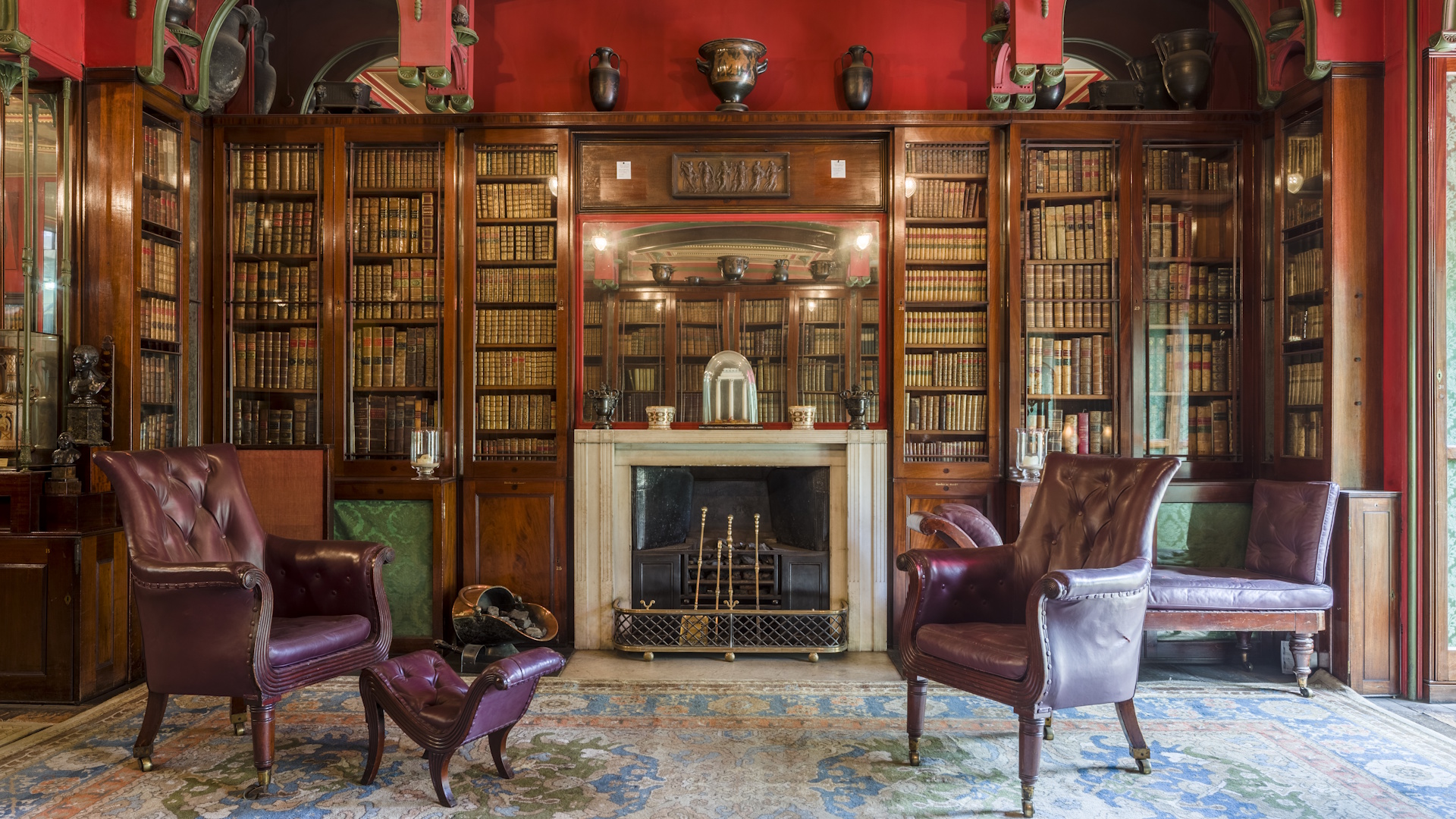 Cosy library room packed with book shelves, a fireplace, leather armchairs, red walls and a big colourful carpet.