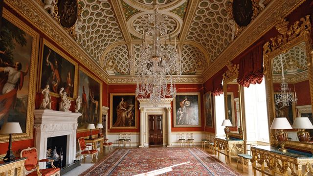 A view along the length of The Great Room in Spencer House, with red walls, framed portraits, chandeliers and a large rug.