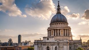 The exterior of St Paul's Cathedral, with its iconic grey-coloured dome, against a bright sky with the sun shining through clouds.