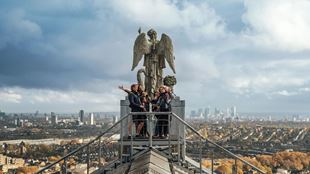 A group standing on the summit platform of Alexandra Palace with the angel statue and London skyline in the distance.