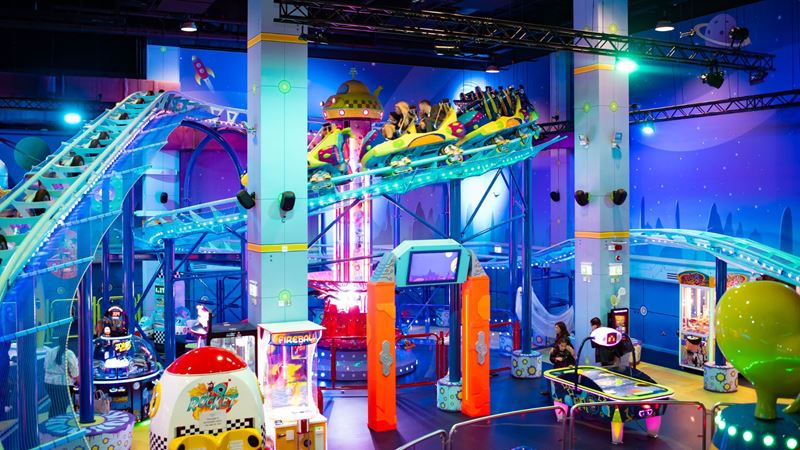 Brightly lit indoor amusement park with roller coaster rides, arcade games, and space‑themed décor.