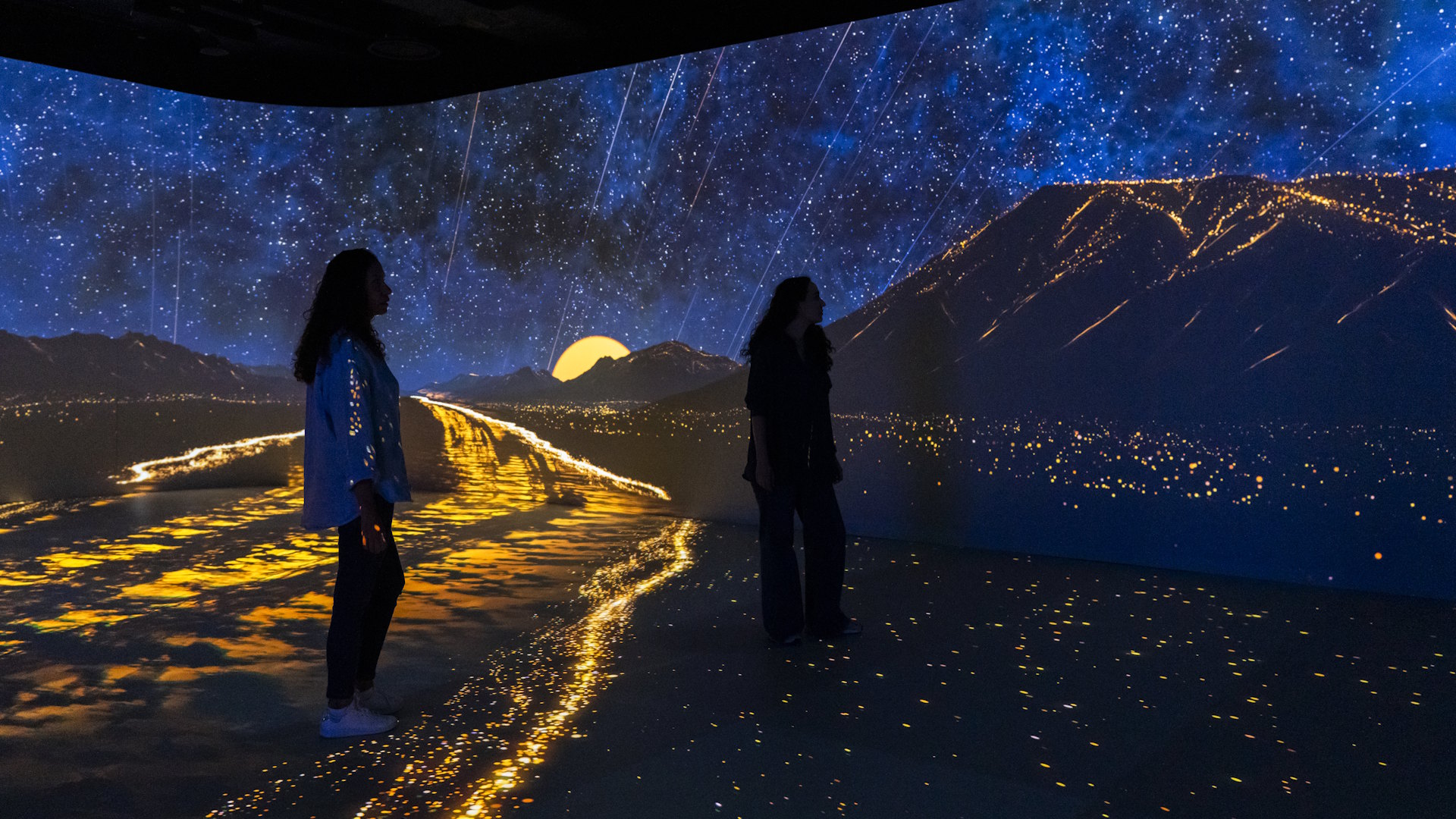 Two female visitors surrounded by blue and yellow light in immersive show depicting a mountain landscape at night.