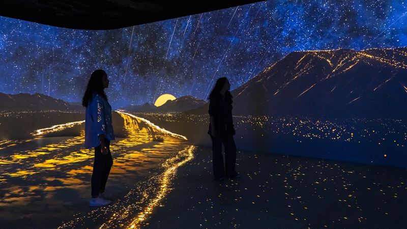 Travel back to Ancient Egypt and explore landscapes illuminated by immersive light and projection mapping at Timewalk. Image credit: Sandra Ciampone. Image courtesy of DEM Museums. Two female visitors surrounded by blue and yellow light in immersive show depicting a mountain landscape at night.