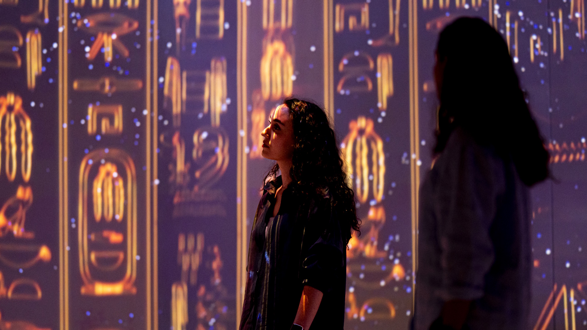 Two female visitors observe ancient Egyptian hieroglyphs projected onto the walls at an immersive exhibition.