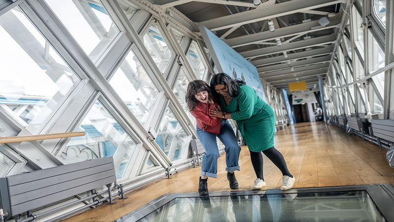 Enjoy panoramic views of the city from the Glass Floors at Tower Bridge. Credit: Michael Barrow. Image courtesy of London & Partners. Two women are holding each other as they are about to walk over the glass walkway at Tower Bridge in London.