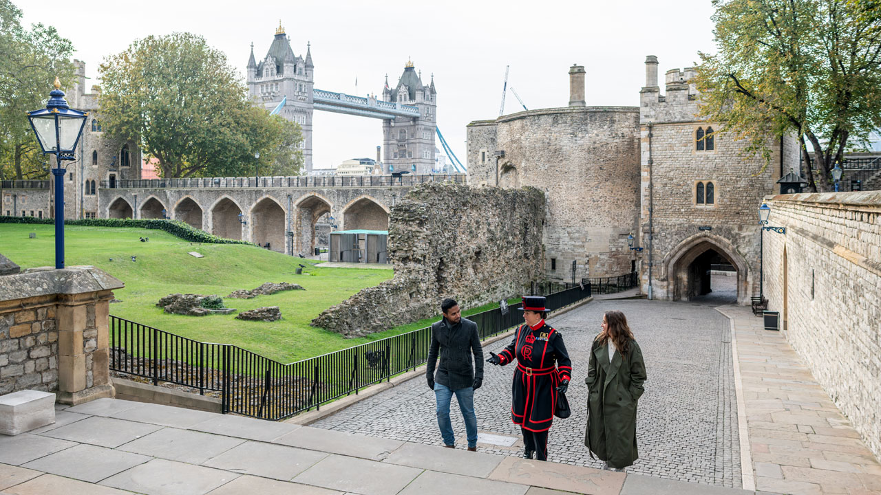 Explorez la Tour de Londres et apprenez-en plus sur ses différentes au fil des ans, guidé par un Beefeater. ©London & Partners/Michael Barrow Un homme et une femme se promènent à l'intérieur de la Tour de Londres lors d'une visite guidée accompagnés d'un garde Beefeater.