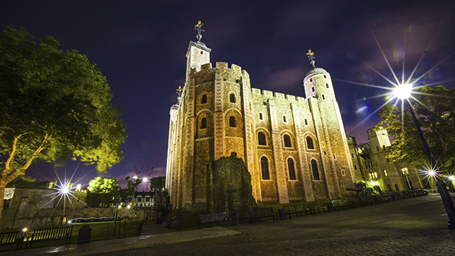 The Tower of London, bei Nacht mit gelben Licht beleuchtet. Ein schwach beleuchteter Baum auf der linken Seite, ein Laternenpfahl auf der rechten Seite und ein violetter, dunkler Himmel erzeugen eine gespenstische Atmosphäre.