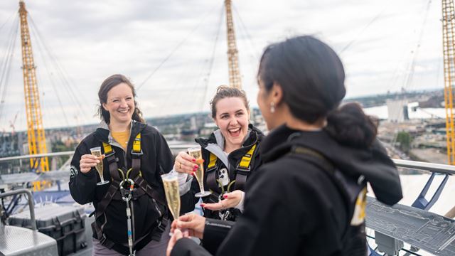 Three climbers wearing safety harnesses share champagne at the top of The O2 in London.