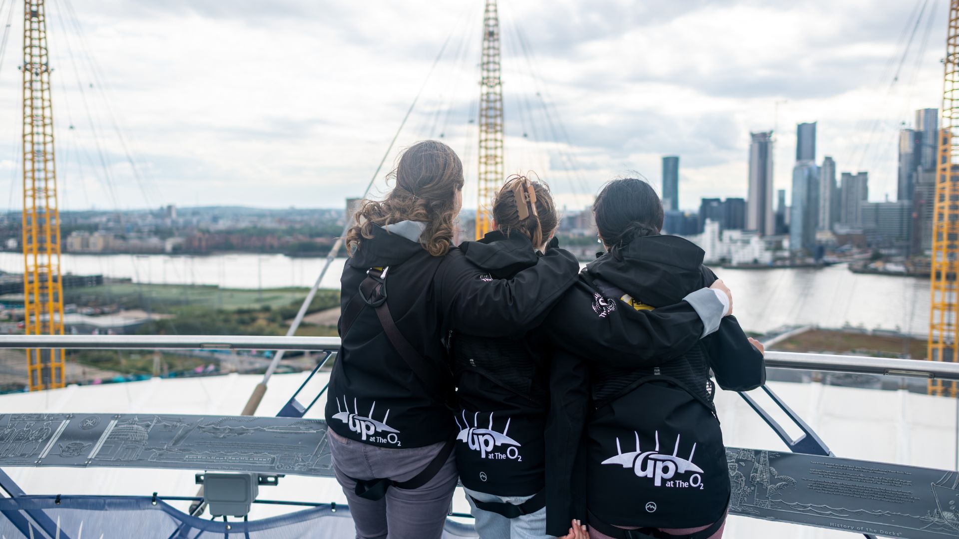 Ein denkwürdiger Moment – Freunde umarmen sich, während sie von der Aussichtsplattform im Up at The O2 die Skyline bewundern. Bild mit Genehmigung von London & Partners/Michael Barrow Drei Freunde, die sich umarmen, bewundern den Blick auf die Stadt vom Dach des The O2 in London.