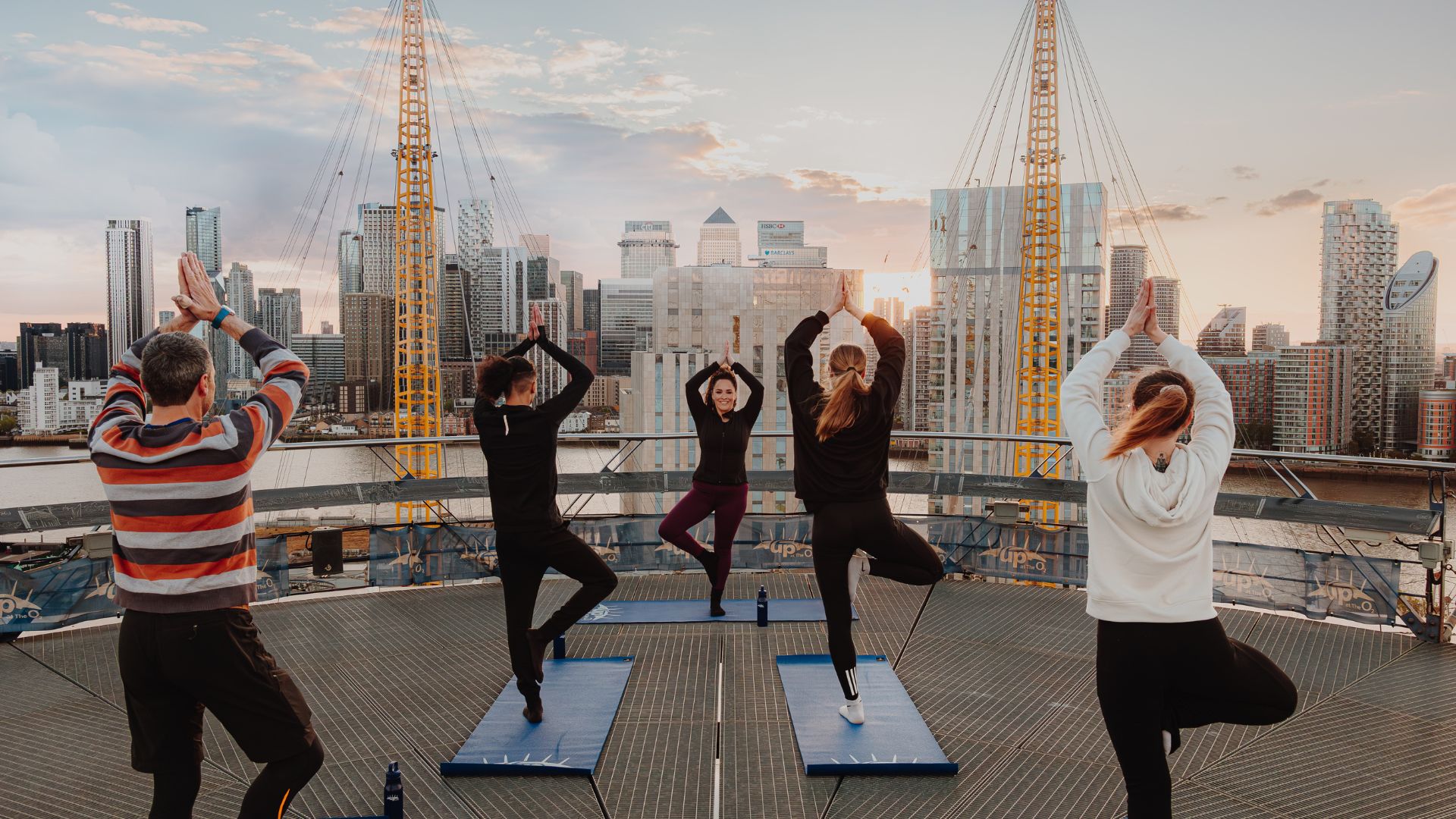 A group of people taking part in a sunset yoga class on top of The O2 in London, with panoramic views of Canary Wharf and the city skyline in the background.