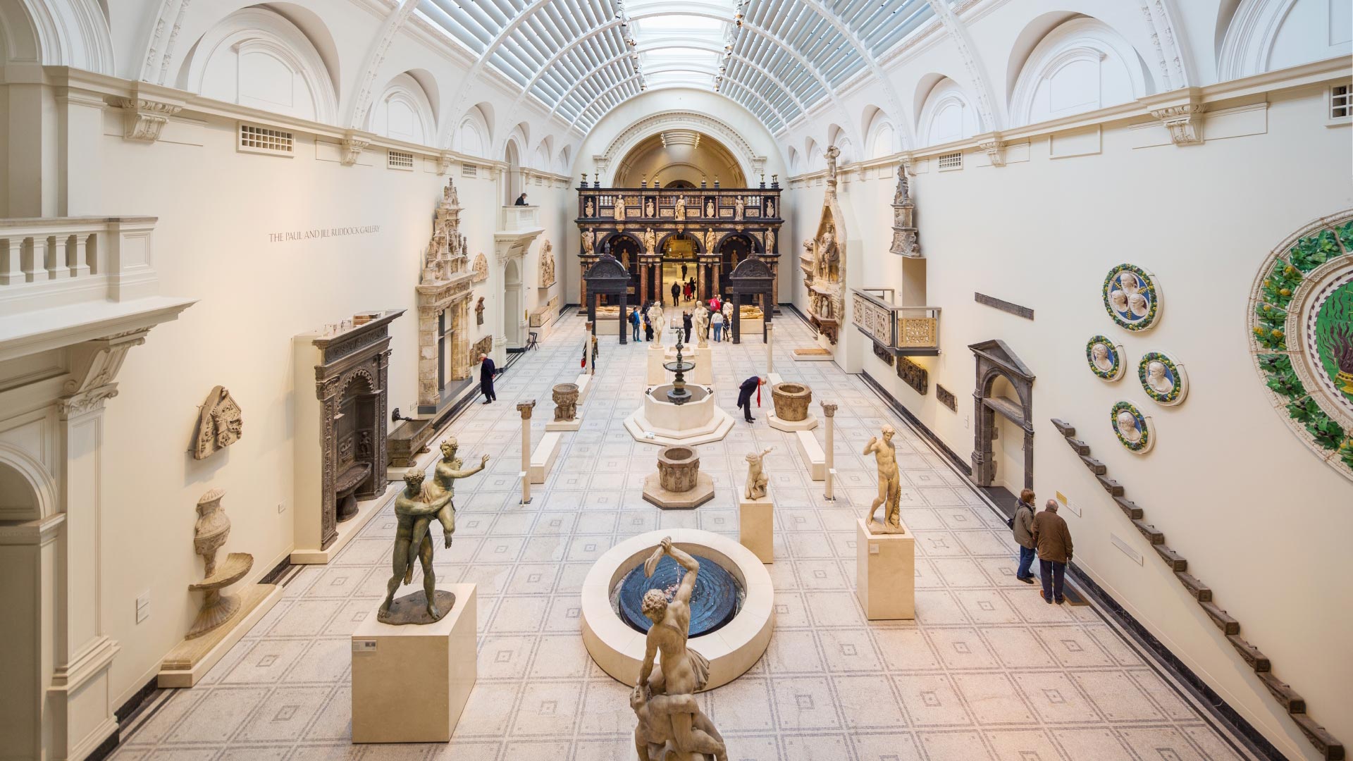 Victoria and Albert Museum. Credit: Jon Reid. Image courtesy of Visit London. A vast light-filled white hall at Victoria and Albert Museum, featuring sculptures and statues on pedestals, artefacts on walls and a curved, glass roof.