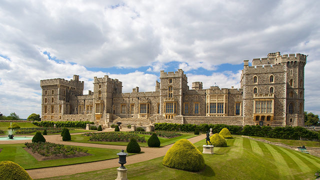L'extérieur du château de Windsor par une journée nuageuse mais lumineuse, avec de l'herbe et des buissons bien entretenus au premier plan.