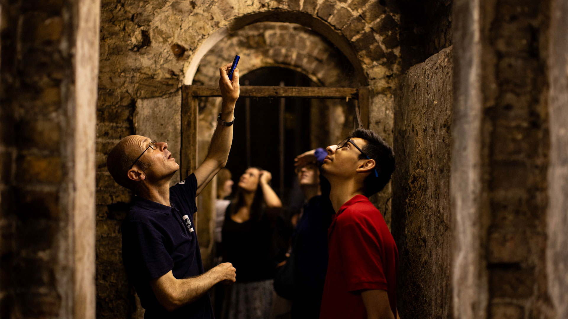 Multiple people standing in underground passageway shining a torch above them.