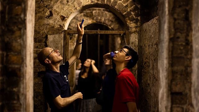 Multiple people standing in underground passageway shining a torch above them. 
