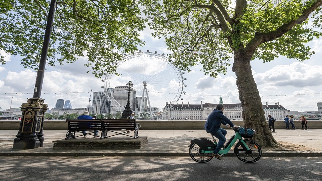 low angle shot of the London Eye from across the water at Embankment, with people strolling past and a man cycling past