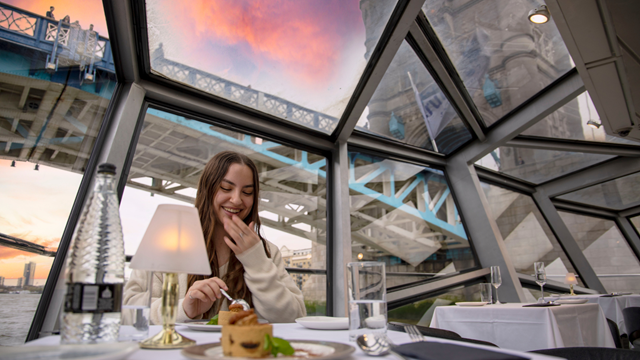 A girl sits at a table on a river cruise with a meal and champagne as the boat rides underneath tower bridge.