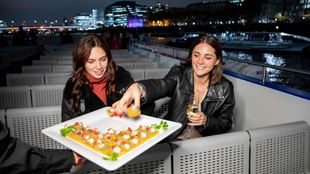 Two people sat on chairs outside on a river cruise taking canapes off a plate.