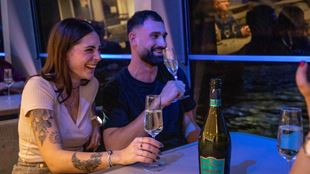 Two people with glasses of presecco are laughing as they look at views of london on a cruise.