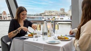 Two people sit at a table with plates of food and glasses of champagne on a city cruises river thames cruise in london.