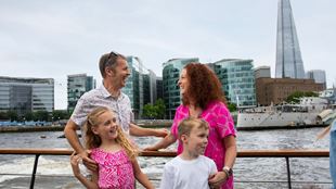 A man in a light shirt stands with a woman and a girl in pink with a young boy in a white shirt as they spot sights on board a river thames cruise boat.