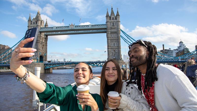 Snap a selfie with London's landmarks on a river Thames cruise. Image courtesy of City Cruises. Three people holding coffees take a selfie in front of Tower Bridge on a river thames cruise boat.