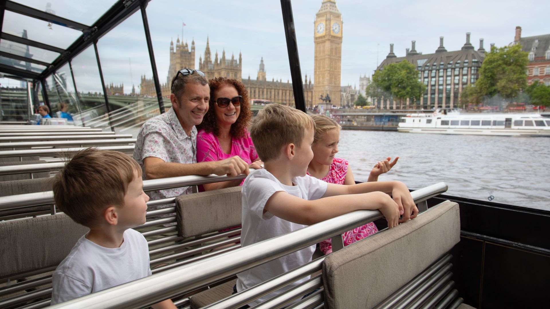 A family of four sit on benches on aboard a river Thames cruise looking out at Big Ben.