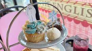 Close-up of an afternoon tea stand featuring a cupcake decorated with The Snowman topper, a snowflake pick, and assorted sweet treats on a bus tour.