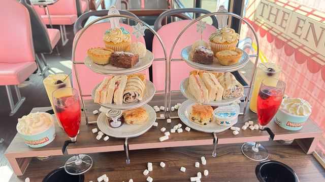 Close-up of an afternoon tea spread on The Snowman-themed bus, featuring sandwiches, scones, cupcakes, and a glass of red sparkling drink.