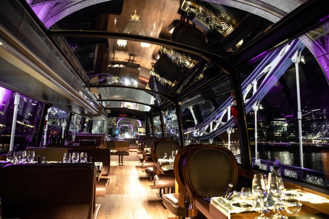 Ein kulinarischer Reisebus mit Sitzplätzen und Tischen, die mit Tellern, Gläsern und Besteck unter einer Glasdecke versehen sind.