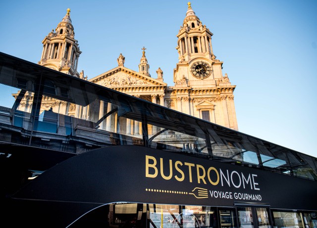 Montez à bord de Bustronome pour une visite mémorable de Londres en bus. Avec l'aimable autorisation de Bustronome. Crédit photo : Bobbymakepeace. Gros plan sur le bus à impérial noir Bustronome se trouvant devant une église par un temps ensoleillé.