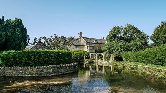 An image of Bourton-on-the-Water showing the river and green bushes surrounding it with a cottage in the back.