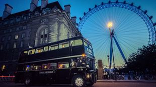 A photo of a black Ghost Bus driving London's streets after dark, with the London Eye in the background.