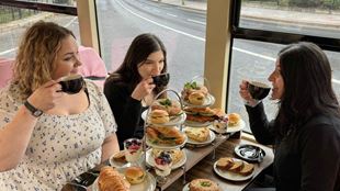A table set for a brunch experience on a bus, featuring a multi‑tiered stand filled with sandwiches, pastries, and berry-topped yogurt parfaits. Several people sit around the table holding cups of hot drinks, with plates of food arranged in front of them.