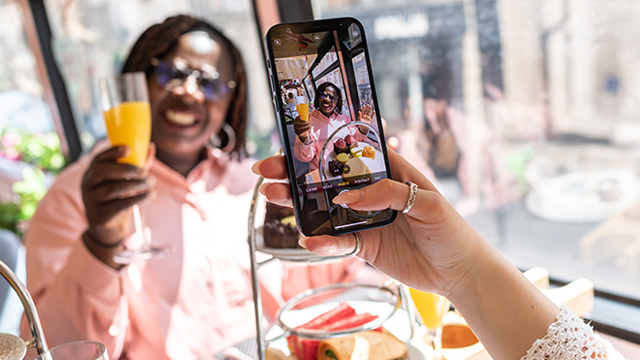 A woman is taking a photo on her smartphone of her friend holding a mimosa during the afternoon tea bus tour of London.