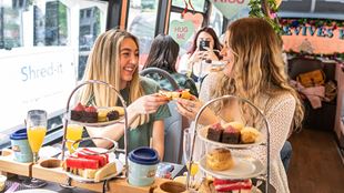 Two women are shown enjoying an afternoon tea aboard a panoramic bus tour of London and are cheering with scones while laughing together.