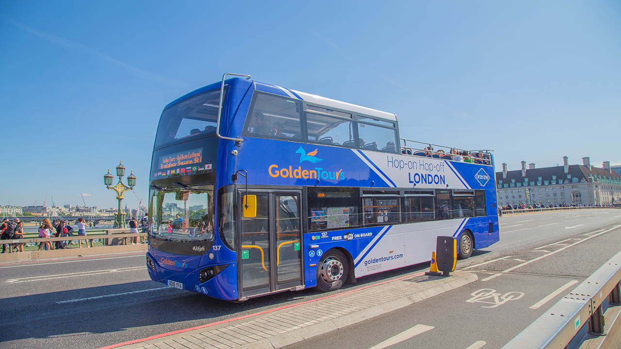 A blue and white double decker sightseeing bus tour is driving past the busy streets of London. 
