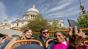 Three people take a selfie from the top of the Hop-On Hop-Off open-top bus, with St Paul's Cathedral visible in the background.