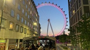 An open top bus filled with people driving towards the London Eye lit up in pink in the evening