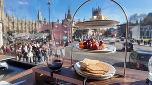 Cheese, grapes, crackers and a glass of red wine on a wooden table by the top-floor window of a double-decker bus overlooking Westminster on a sunny day with blue sky.