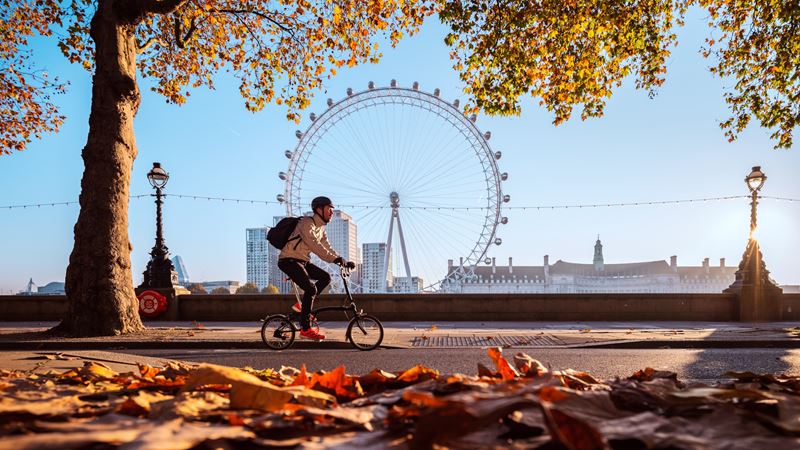 Cycle along the river Thames to discover the city's hidden gems. Credit: Antoine Buchet. Image courtesy of visitlondon.com. Man cycling along the river Thames on a clear autumn day with the London Eye in the background and autumn leaves in the foreground.