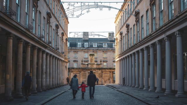 Three people walk through the old streets of bath where christmas lights hang above their heads.