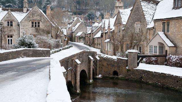 A snowy street and bridge over flowing water with honey coloured stone cottages on each side of the road