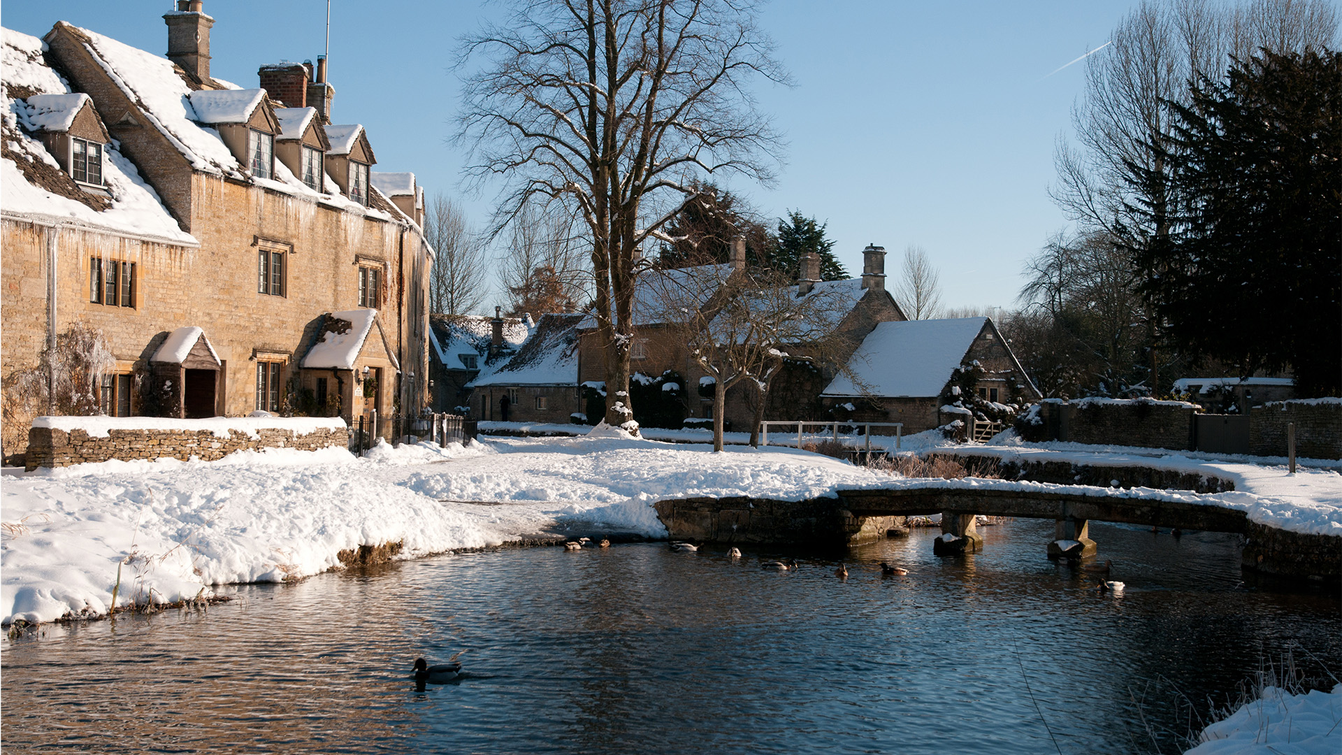 Visit the charming villages of the Cotswolds during the festive season. Image courtesy of Golden Tours/Shutterstock. A river with a bridge over it and some cottages covered in snow.