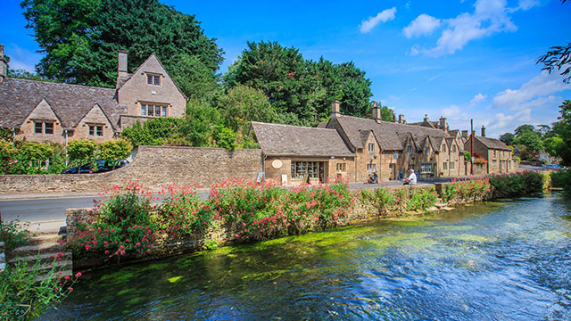 Une rivière, au premier plan, traverse un joli village des Cotswolds, avec, à l'arrière-plan, une petite route et des maisons construites avec la pierre locale de couleur dorée.