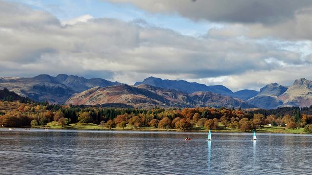 Sailboats on Lake Windermere with mountains and trees in the background, on a cloudy day.