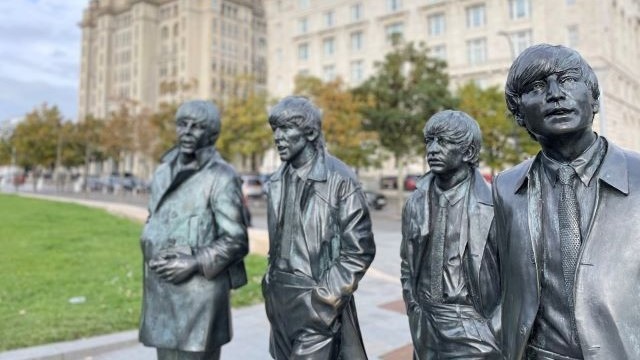 The Fab Four statue in Liverpool, with grass, blue sky and a building in the background.