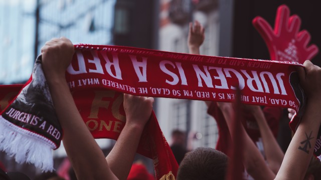 Liverpool football supporters holding red scarves in the air with 'Jurgen is a red' slogan written across the scarves.