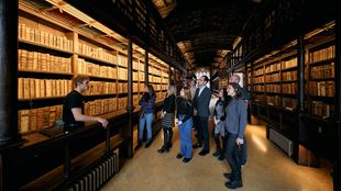 A guided group is standing in front of the book shelfs within Duke Humfrey's Library at Bodleian Library and the guide is telling them something.