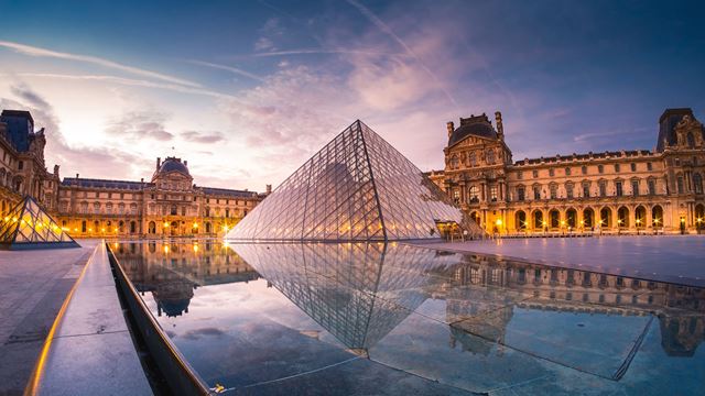 An evening view of the Louvre's iconic pyramid from across a water feature, with the facade of the museum illuminated.