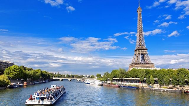 A view along the river Seine on a sunny day, with a river boat in the foreground and the Eiffel Tower in the background.